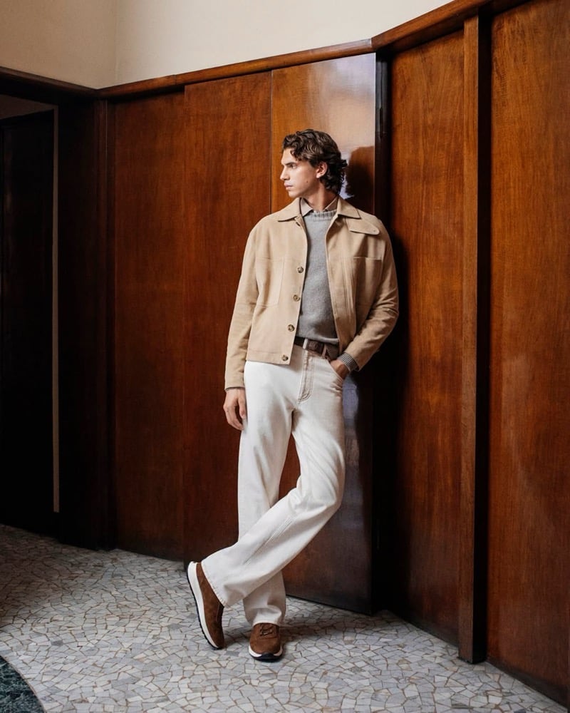 Hedi Ben Tekaya leans against a wood-paneled wall in a camel suede jacket, gray sweater, and jeans, wearing brown suede sneakers