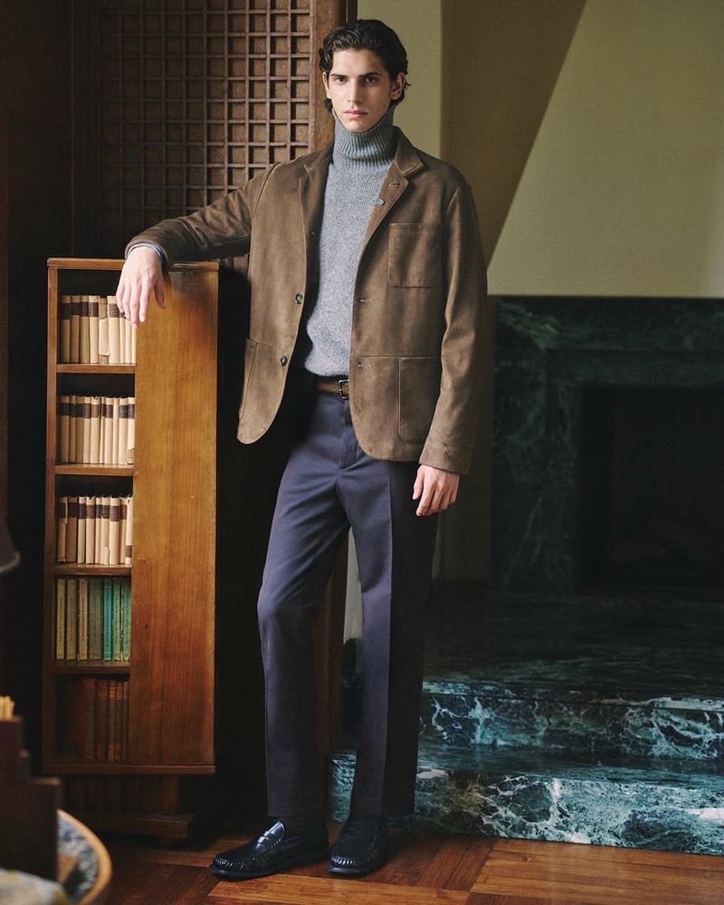 Hedi Ben Tekaya wears a brown suede blazer over a gray rollneck with tailored trousers and leather loafers beside a bookcase for Tod's fall-winter 2026 campaign