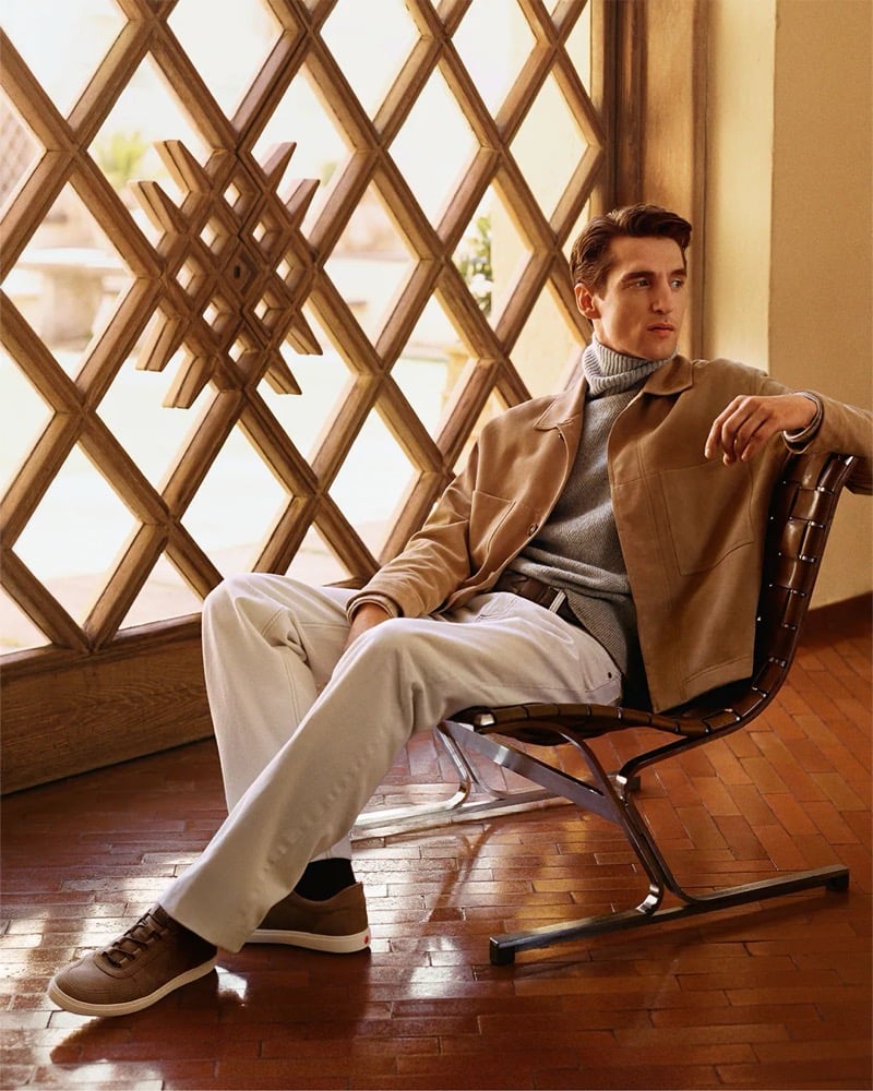 Anatol Modzelewski sits in a leather sling chair in a tan suede overshirt, gray rollneck, and suede sneakers by a latticed window for Tod's fall-winter 2026 campaign
