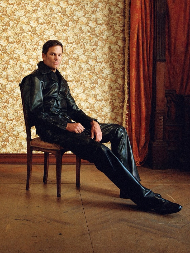 Tom Brady sits on a wooden chair against floral wallpaper in a black leather jacket and trousers for Gucci's Primavera campaign