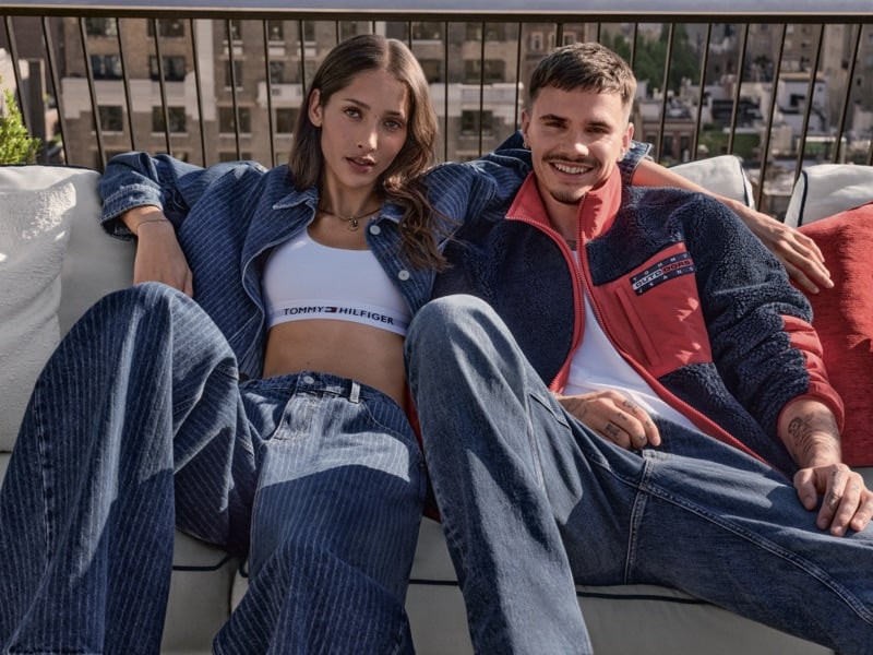 Romeo Beckham sits on a rooftop couch in a navy and red fleece jacket and jeans beside a model in a pinstriped denim jacket and white logo bra top