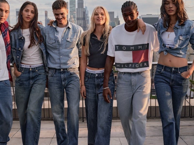 Romeo Beckham, Yasmin Wijnaldum, and four cast members stand in denim jackets and jeans, with a white Tommy Hilfiger logo t-shirt among the looks, city skyline behind them in daylight