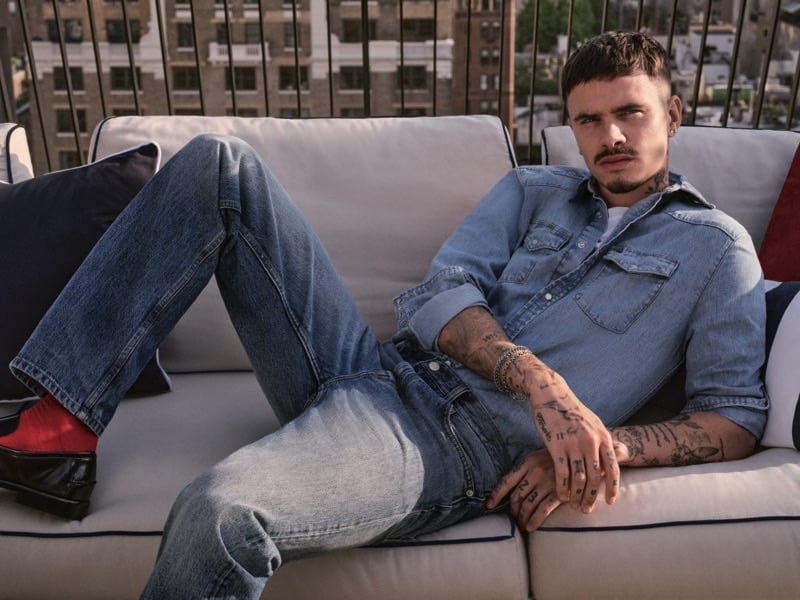 Romeo Beckham lies across a rooftop couch in a denim shirt and jeans with red socks and black loafers, New York City visible through the railing