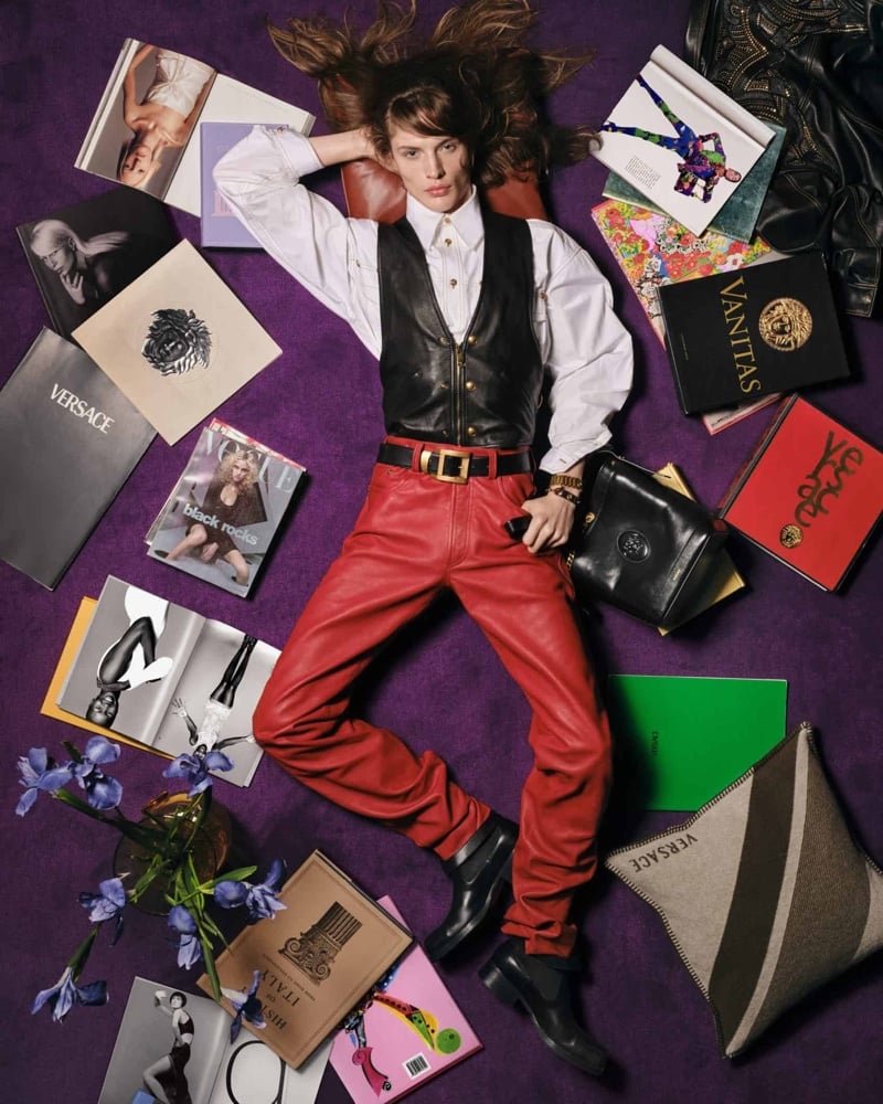 Wiley Rhoads lies across a purple backdrop scattered with Versace magazines and books, wearing a black leather vest, white shirt, and red leather trousers, with a black leather shoulder bag and Chelsea boots