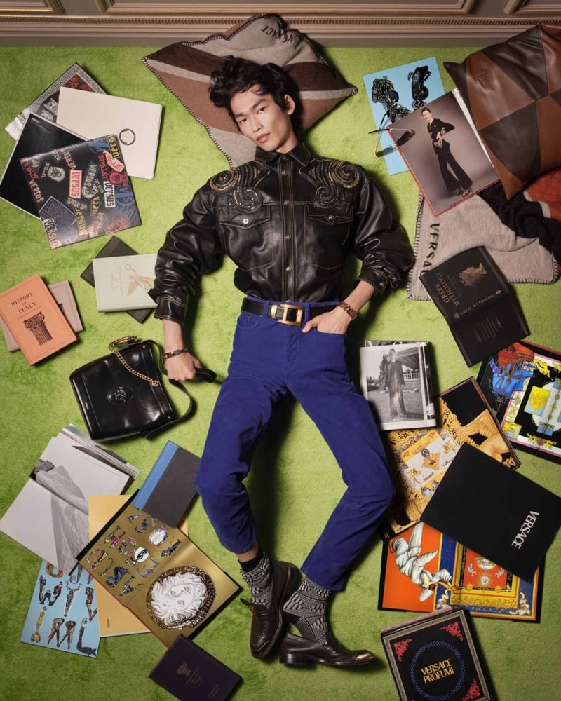 Bai lies across a green backdrop surrounded by Versace magazines and objects, wearing an embroidered black leather shirt, blue trousers, a gold Medusa belt, and patterned socks with black loafers