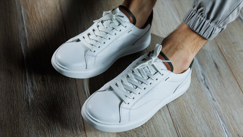 A close-up of white leather sneakers on a person's feet standing on a wood floor