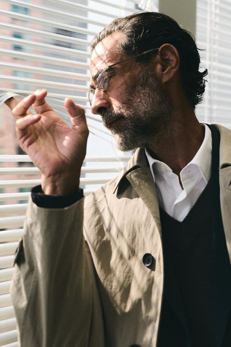 John Armstrong wears a tan trench coat while adjusting window blinds for Zara's back to work 2026 campaign