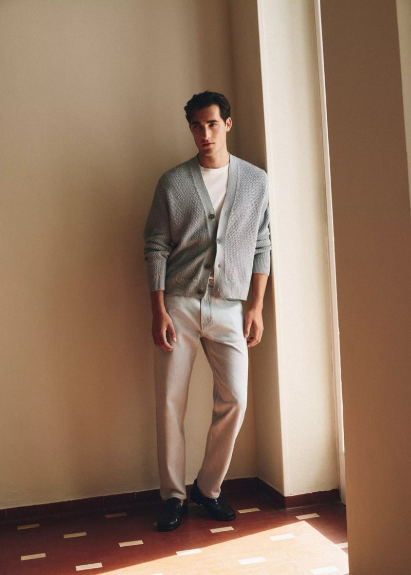 Adam Sattrup stands in a doorway in a gray cardigan over a tee with relaxed pale wash jeans and black loafers, for Mango's Superior Denim collection