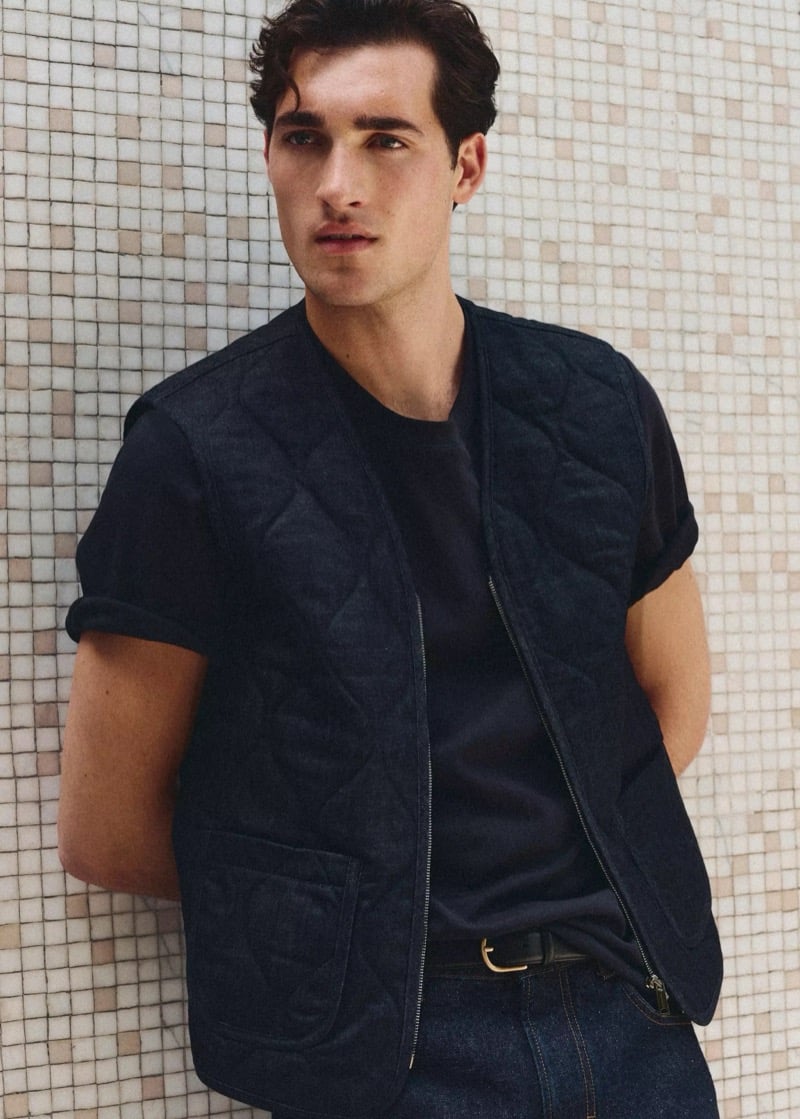 Adam Sattrup wears a dark quilted gilet over a crewneck tee against a mosaic tile wall, for Mango's Superior Denim collection