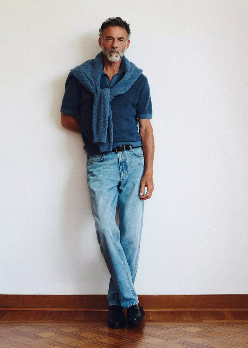 Alain Gossuin leans against a plain wall with a cable-knit sweater tied over a polo shirt and pale wash jeans, for Mango's Superior Denim collection