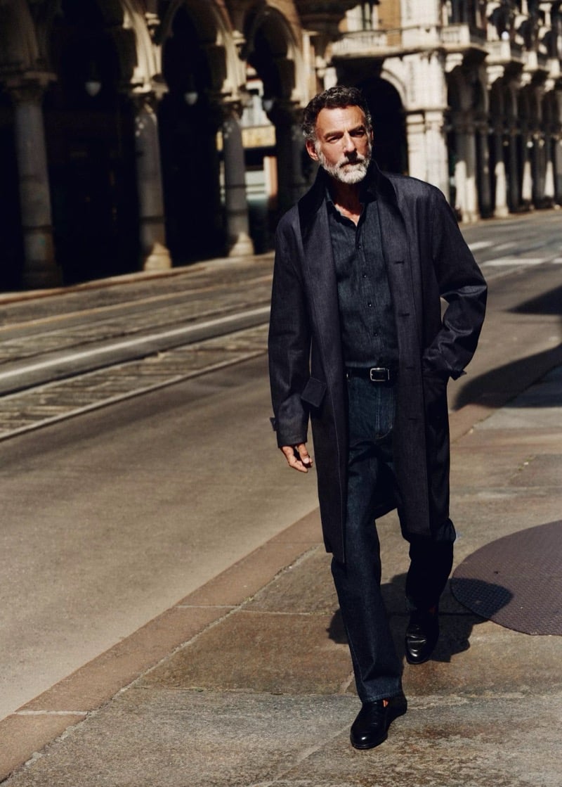 Alain Gossuin walks beneath a colonnade in a long indigo denim trench coat over a button-front shirt with relaxed dark jeans, for Mango's Superior Denim collection