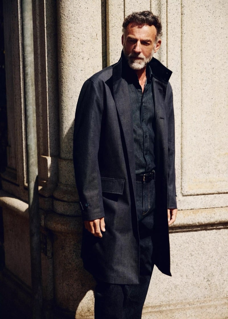 Alain Gossuin stands against a ribbed stone wall in a denim trench coat with a stand-up collar over a button-front shirt, for Mango's Superior Denim collection