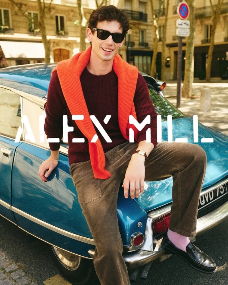 Tom Hughes sits on the hood of a vintage blue Citroën in sunglasses, wearing a burgundy washed cashmere sweater with an orange ribbed knit over his shoulders, brown corduroy trousers, pink socks, and black loafers
