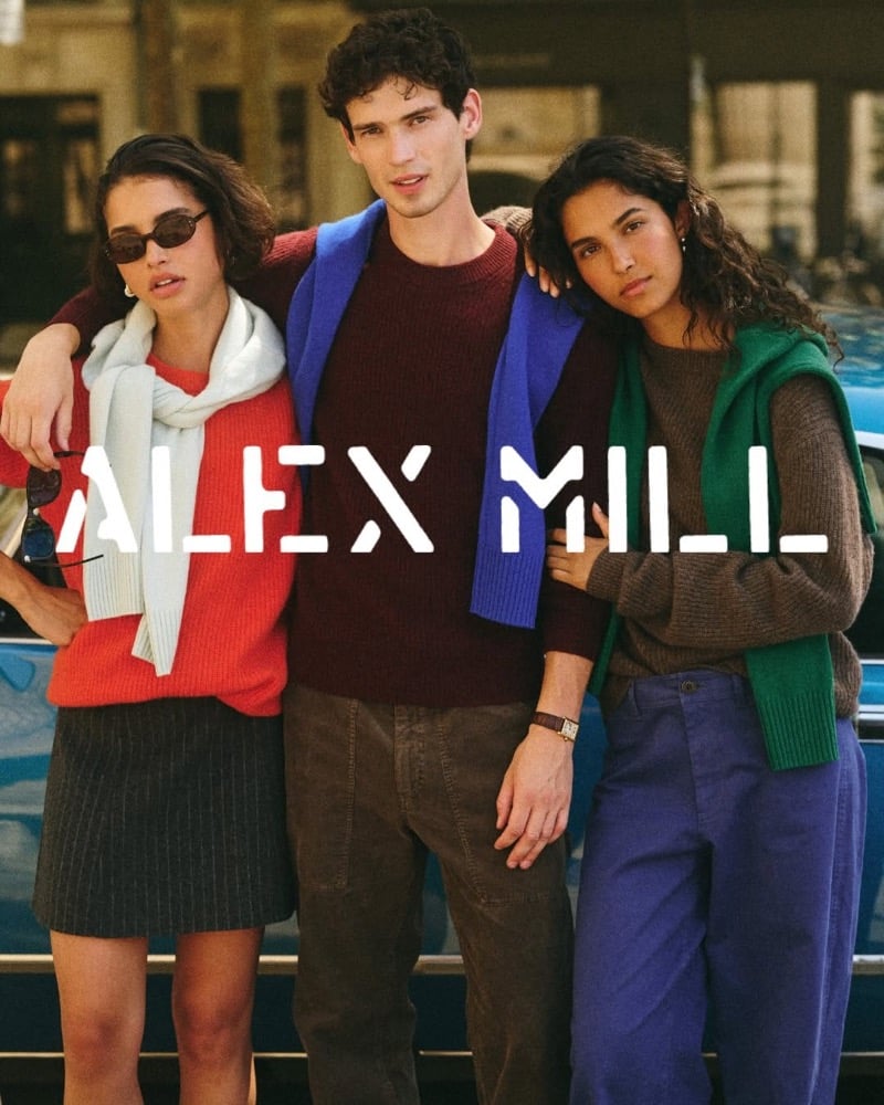 Tom Hughes stands between two female models in front of a blue car, wearing a burgundy crew-neck sweater with a cobalt-blue knit over his shoulders and brown corduroy trousers, the Alex Mill logo set across the image