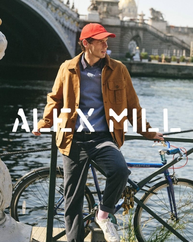 Tom Hughes leans against railings by the Seine with a bicycle, wearing a red baseball cap, a tan moleskin jacket, a stormy sea sweatshirt over a collared shirt, and dark trousers with pink socks and sneakers