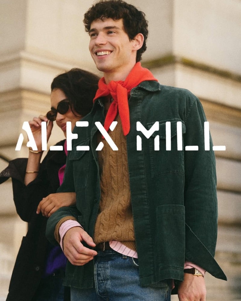 Tom Hughes smiles outdoors in a green corduroy chore jacket worn open over a tan cable-knit sweater and a pink striped shirt, with a red neckerchief and blue jeans, a woman in sunglasses beside him