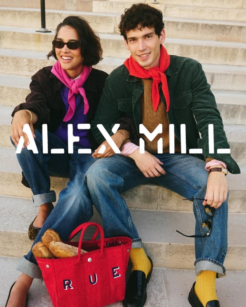 Tom Hughes and a woman sit close together on stone steps, each in a knotted neckerchief; he wears a green corduroy chore jacket over a tan cable-knit sweater with cuffed jeans, yellow socks, and black loafers, a red tote of baguettes on the step below