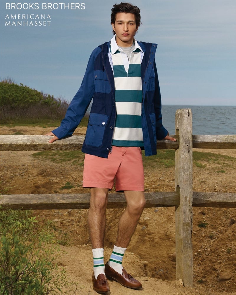 Ruben Bilan-Carroll leans against a weathered wooden fence by the ocean, wearing a blue two-tone hooded anorak over a green-and-white striped rugby polo with coral chino shorts, striped athletic socks, and brown tassel loafers
