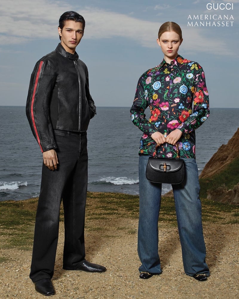 Ruben Bilan-Carroll in a black leather moto jacket with a red racing stripe and dark jeans beside Ava Shipp in a dark floral-print shirt, jeans, and a small black top-handle bag, standing on a grassy bluff above the sea for Gucci