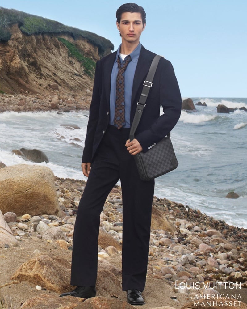 Ruben Bilan-Carroll stands on a rocky Montauk shoreline in a dark single-breasted suit worn with a blue shirt, a brown check-print tie, and a Louis Vuitton monogram messenger bag on a wide strap