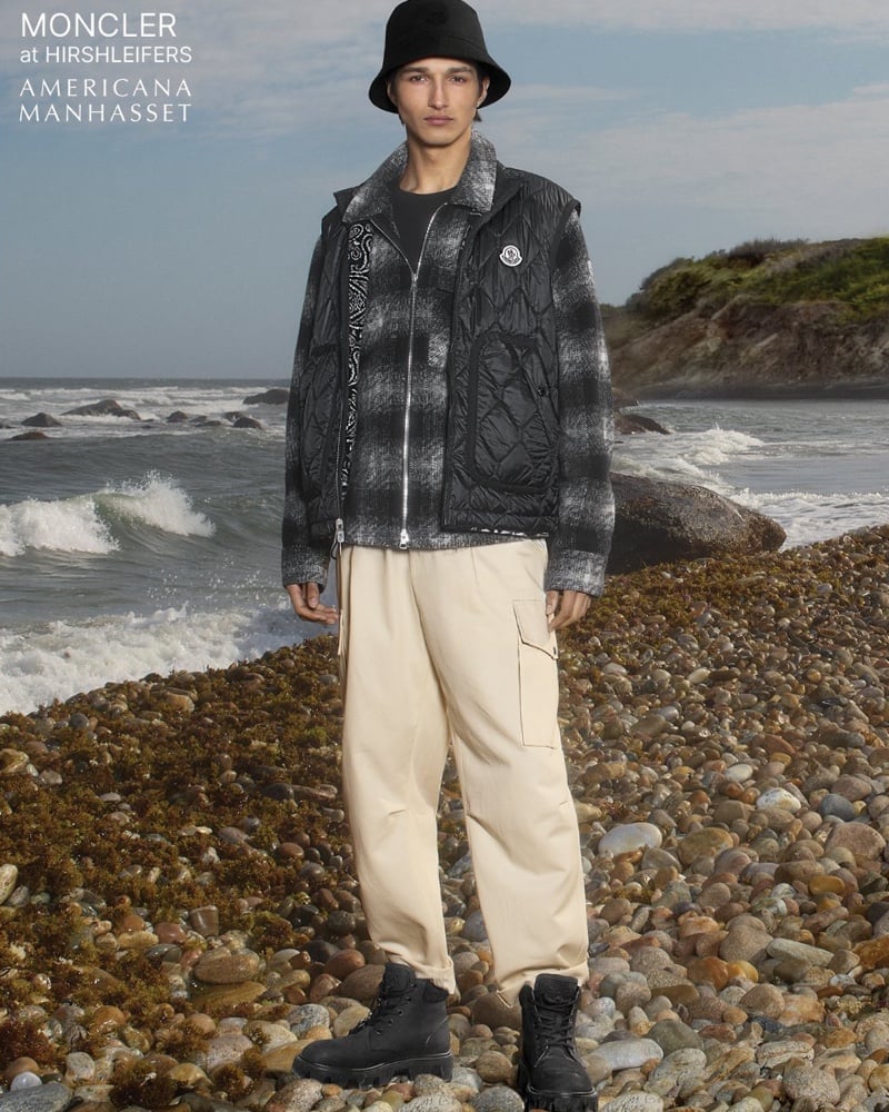 Ruben Bilan-Carroll stands on a pebble beach in a black bucket hat and a plaid flannel shirt jacket layered under a black quilted down vest, with pale wide-leg cargo trousers and black lace-up boots