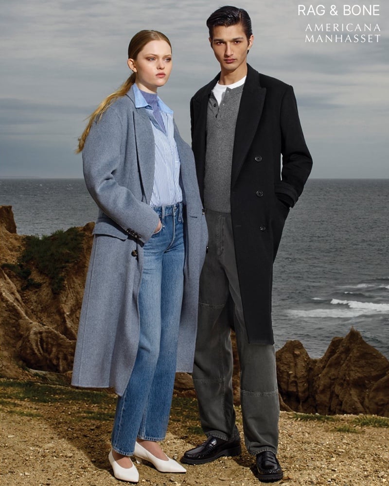 Ava Shipp and Ruben Bilan-Carroll stand together on a bluff above the water; she wears a long soft blue-gray wool coat over a striped shirt and straight-leg jeans with white slingback flats, and he wears a dark double-breasted overcoat over a gray half-zip knit with gray trousers and black loafers
