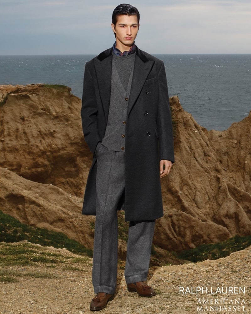 Ruben Bilan-Carroll on an eroded cliff in a charcoal overcoat over a gray V-neck sweater, a checked waistcoat, and Prince of Wales check trousers with brown tassel loafers, wearing Ralph Lauren