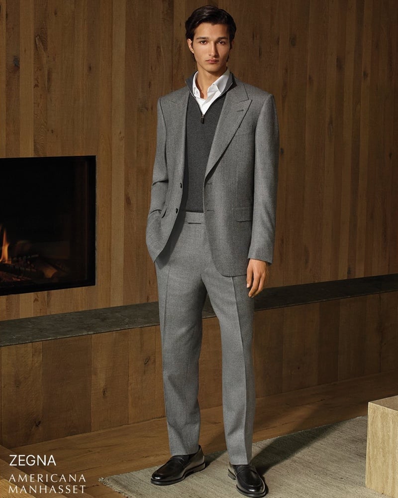 Ruben Bilan-Carroll in a gray tailored suit worn over a charcoal quarter-zip sweater and a white shirt with black loafers, standing by a lit fireplace in a wood-panelled room for Zegna