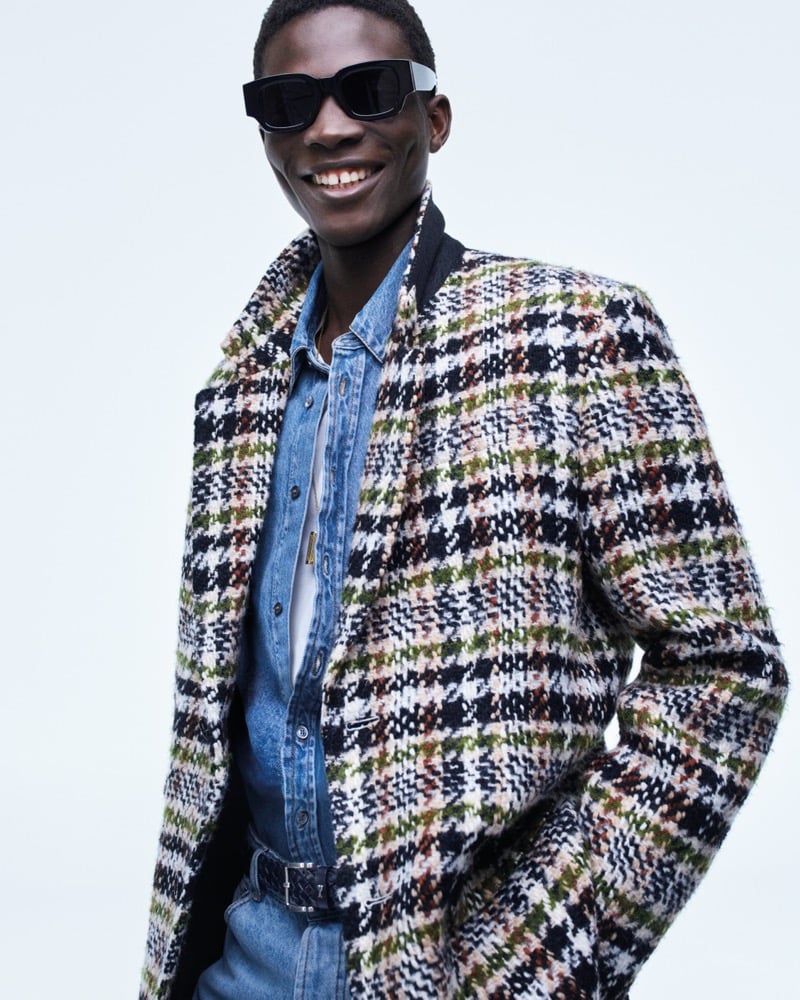 Dara Gueye smiles in black rectangular sunglasses, wearing a black-and-green checked bouclé overcoat over a denim trucker jacket and jeans with a braided belt