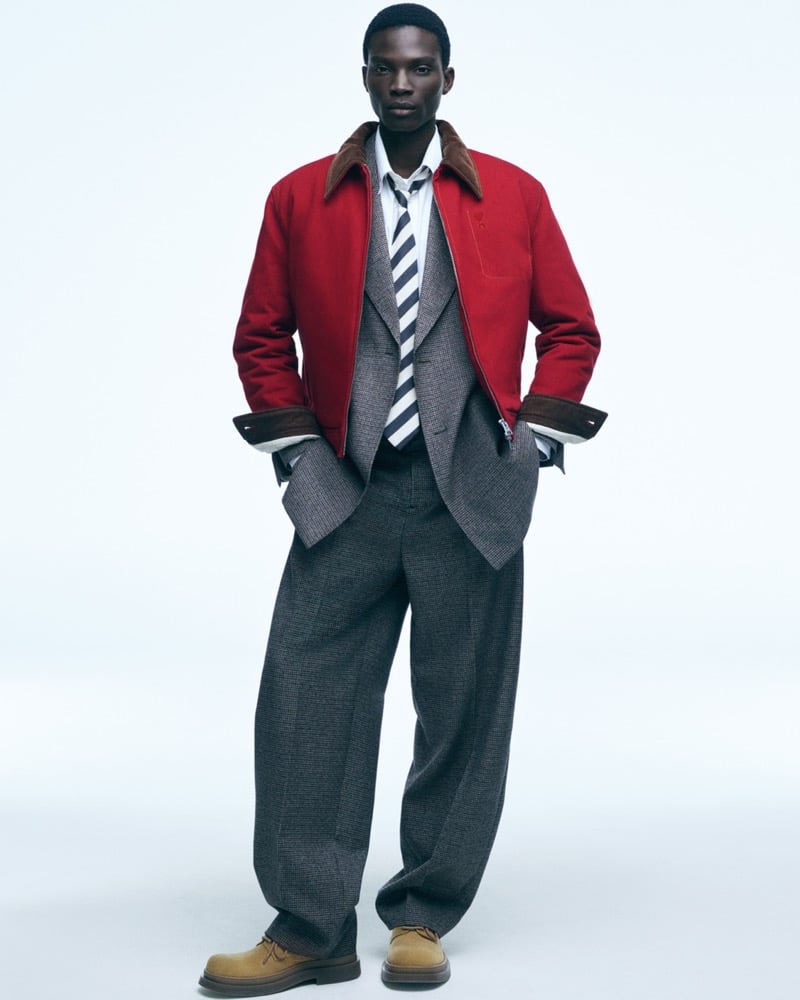 Dara Gueye in a red blouson jacket with a corduroy collar over a grey checked waistcoat and matching wide trousers, a striped tie, and suede desert boots, against a pale backdrop