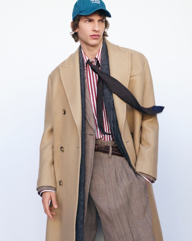 Elliot Belot stands against a pale backdrop in a double-breasted camel overcoat over a gray scarf, a red-striped shirt, and a brown knit tie, with pinstripe pleated trousers, a braided belt, and a teal Ami baseball cap reading For Being a Friend