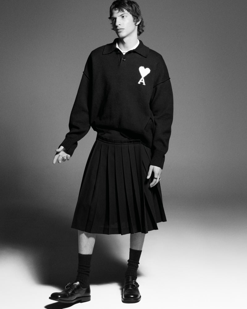 Black and white full-length shot of Elliot Belot in an oversized knit polo shirt with the Ami de Coeur heart logo, a knee-length pleated skirt, ribbed socks, and monk-strap shoes