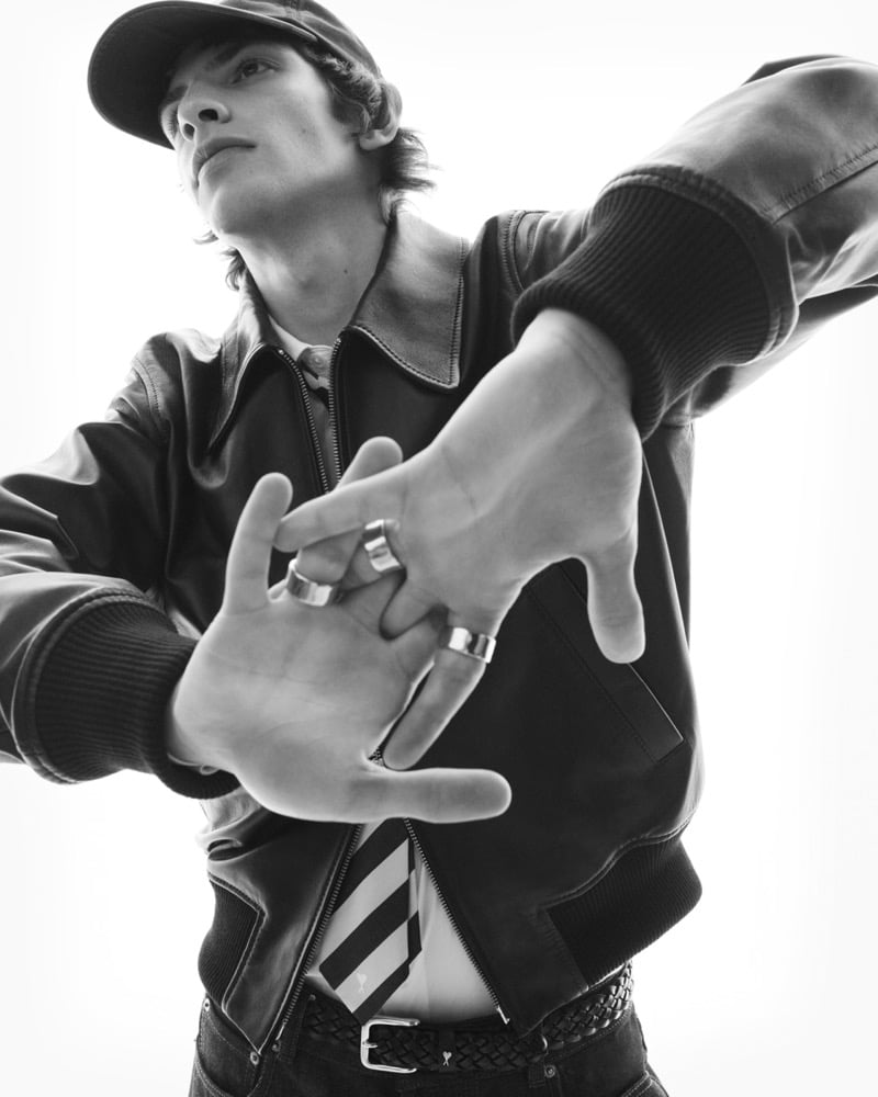 Black and white low-angle shot of Elliot Belot in a leather bomber jacket and cap, stacked silver rings on his fingers as he reaches toward the camera, a striped shirttail and braided belt at the waist