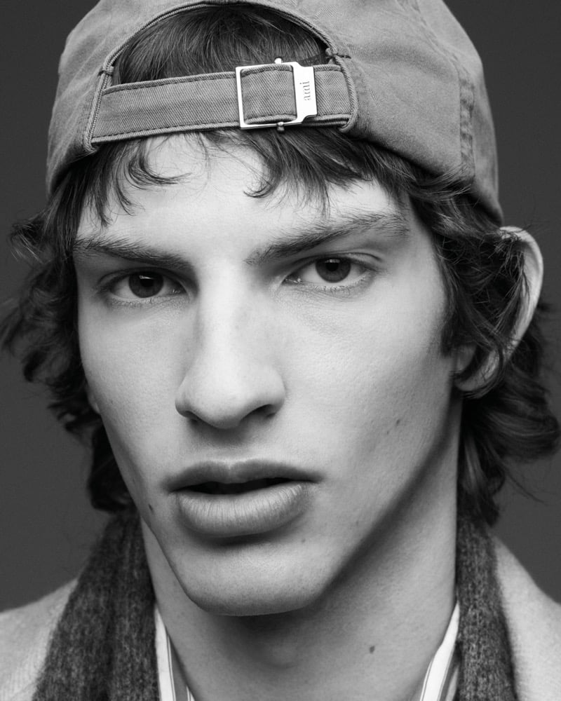 Black and white close-up portrait of Elliot Belot wearing an Ami baseball cap turned backwards, with a marled scarf and striped shirt visible at the neck