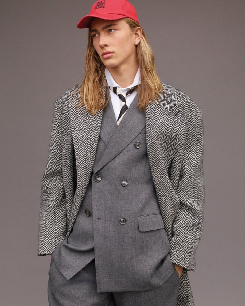 Mack Karpes in a red Ami cap reading Don't Forget to Love, an oversized herringbone tweed coat draped over a grey double-breasted suit with a white shirt and striped tie