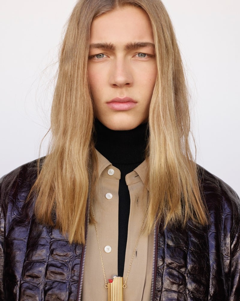 Close-up of Mack Karpes with long straight blond hair, wearing a croc-embossed leather bomber jacket over a tan overshirt and a dark turtleneck, with a slim gold pendant necklace