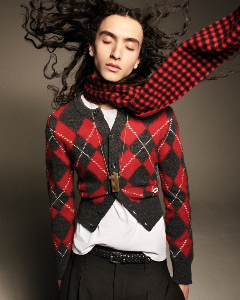Mohamed Benhadda with long curly dark hair blowing across his face, eyes closed, wearing a red and charcoal argyle cardigan over a white T-shirt with a red buffalo-check scarf and a braided belt