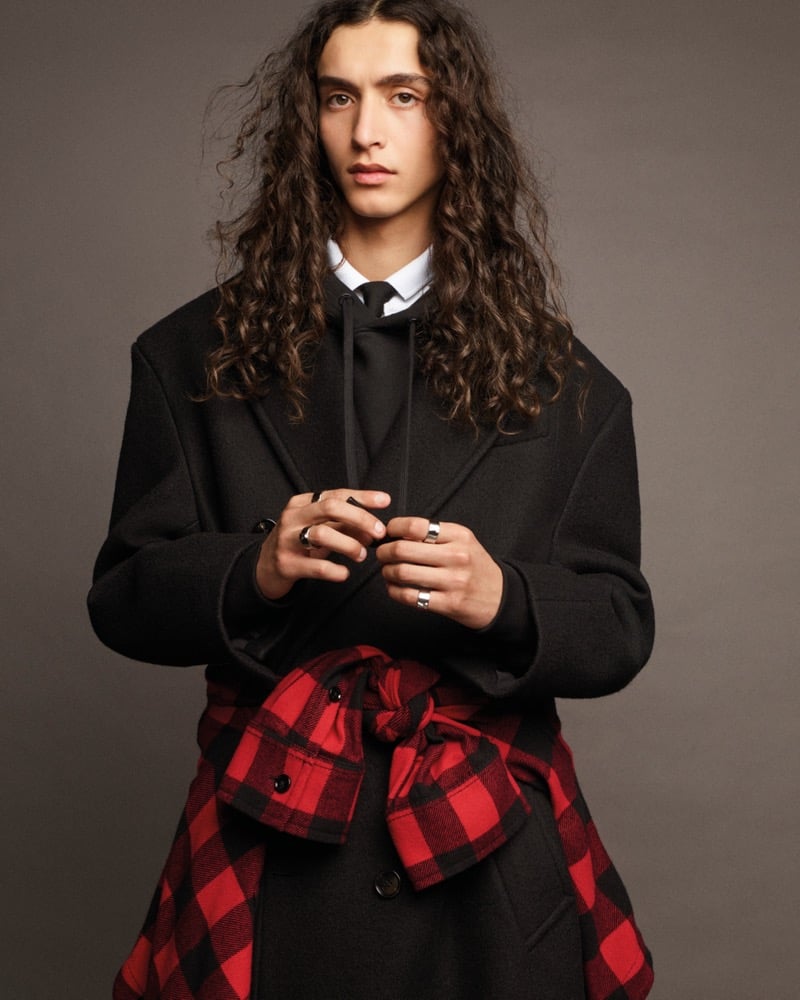 Mohamed Benhadda with long curly hair, wearing a dark tailored overcoat over a hooded top and white shirt with a slim tie, a red buffalo-check shirt knotted around his waist and silver rings on both hands