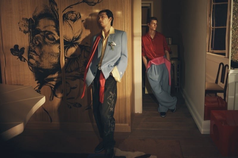 Bai in a light blue blazer with a flower on the lapel and a red scarf, with Roman Lenoir in a red shirt and wide trousers leaning in a doorway behind him