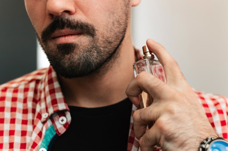 Bearded man in a red gingham shirt spraying fragrance on his neck