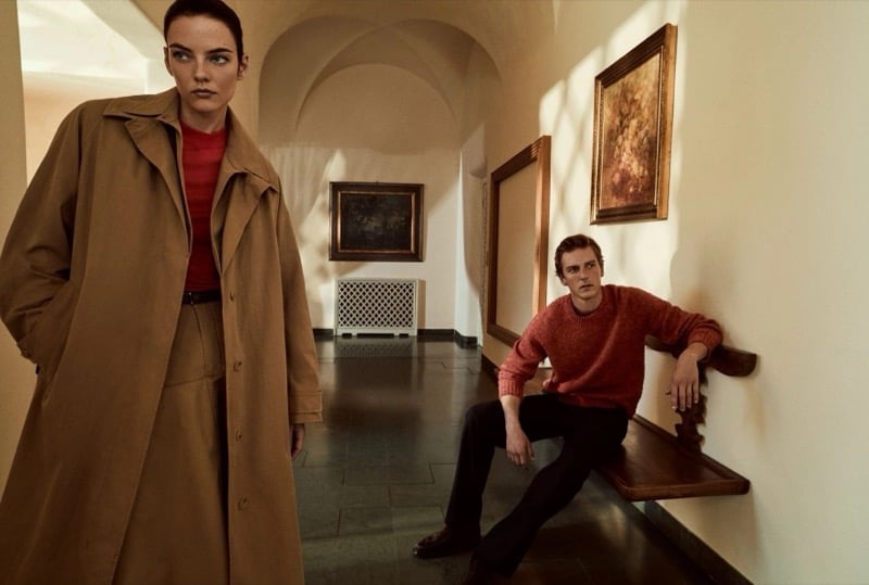 Beauïse Genç in a long trench coat in a vaulted hallway while Quentin Demeester sits on a wooden bench behind her in a knit sweater