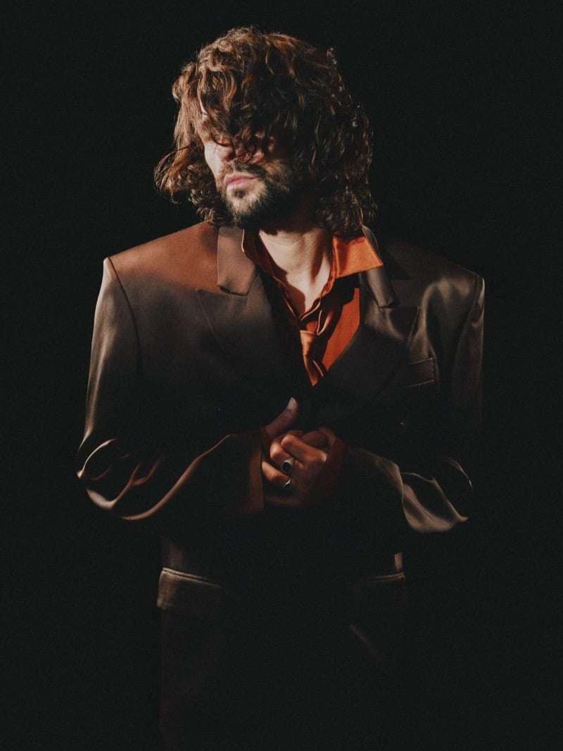 Ben Whishaw in near-darkness pulling a glossy tailored blazer with sharp peak lapels and strong shoulders over an open rust shirt, a silver ring on one hand