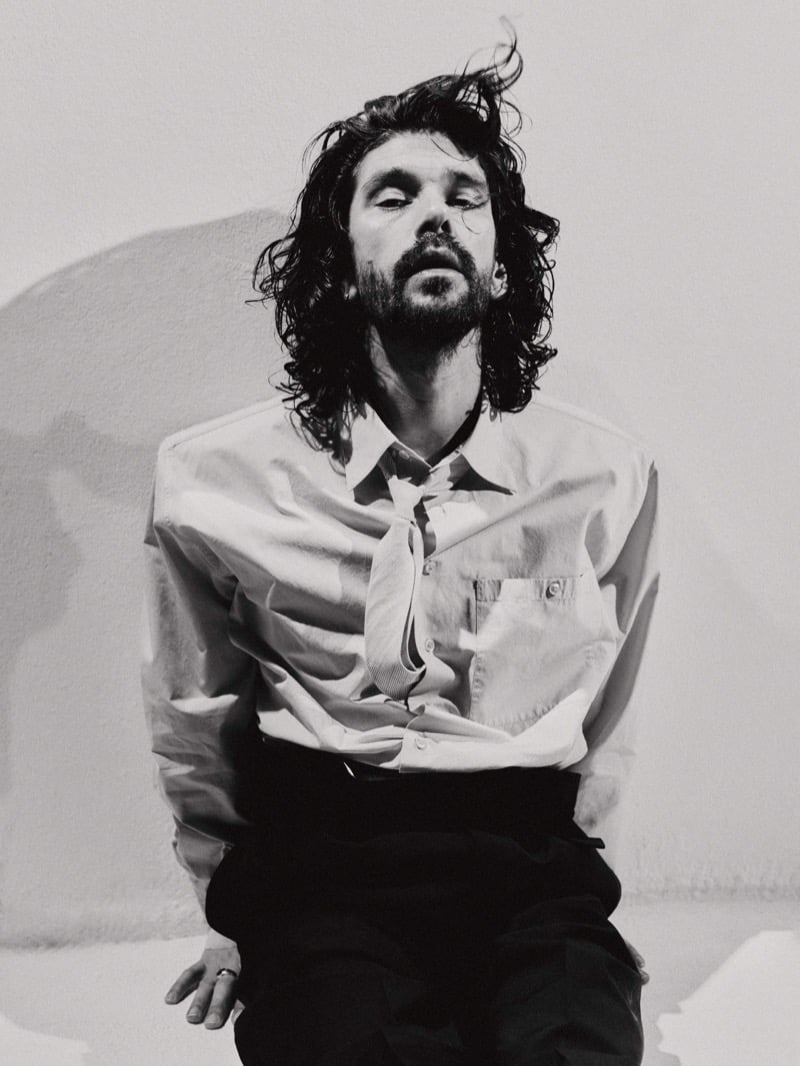 Black-and-white photo of Ben Whishaw seated on a low block, leaning forward in an oversized shirt with structured shoulders and a loosened tie, dark trousers, windblown hair