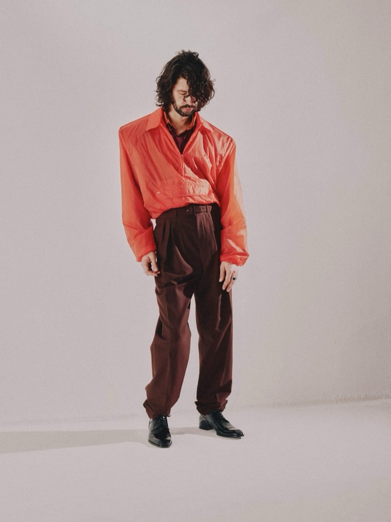 Full-length photo of Ben Whishaw standing with head down in a sheer coral pullover shirt and oxblood high-waisted pleated trousers with turned-up hems and glossy black shoes