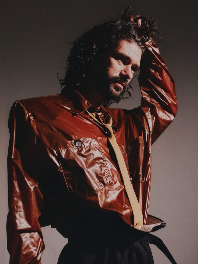 Ben Whishaw raising a hand to his hair in a high-shine metallic copper shirt-jacket with patch pockets and a loosely hanging yellow tie, dark trousers below