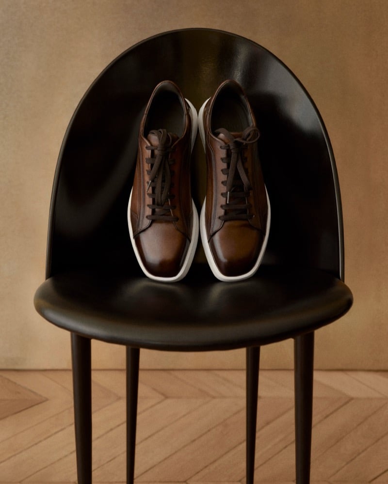 A pair of brown burnished leather Berluti low-top sneakers with white soles placed on a black curved chair above herringbone parquet flooring