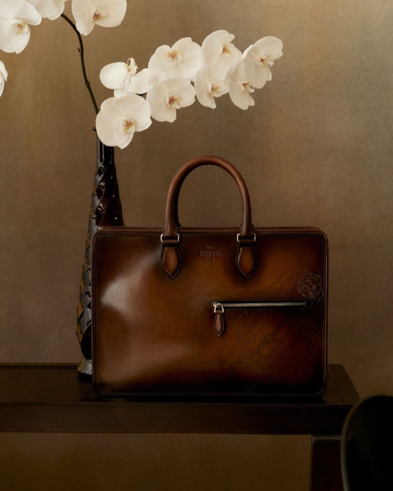 A brown patinated leather Berluti Un Jour briefcase resting on a dark console table beside a stem of white orchids