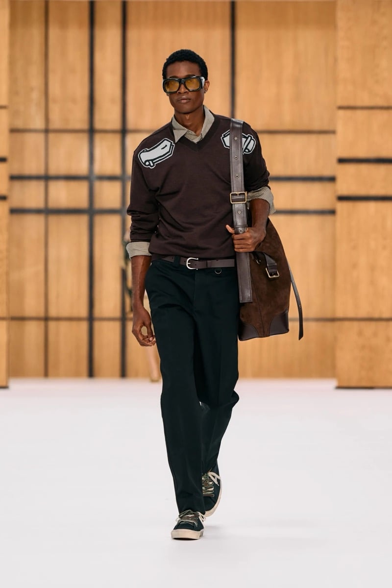 A model in a brown V-neck sweater with appliqué patches, trousers, and a suede shoulder bag, for Burberry's summer 2027 collection
