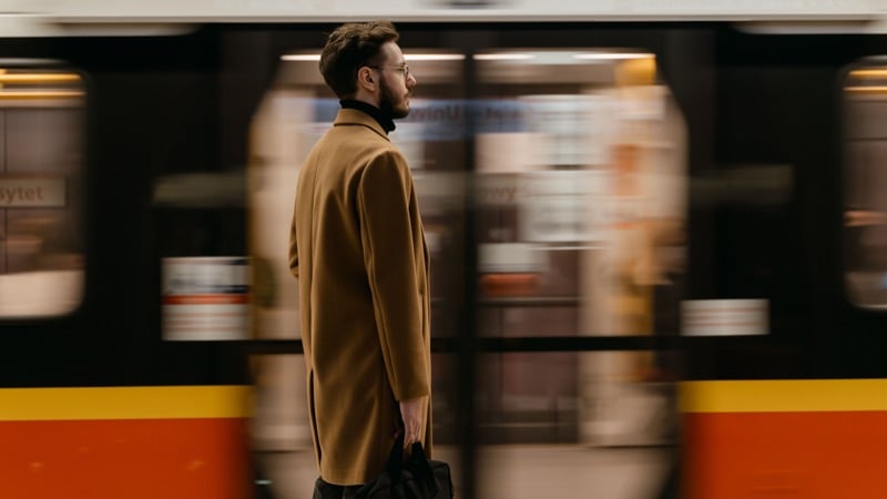 A man in a camel overcoat and black turtleneck waits on a platform with a bag in hand as a train passes in a blur