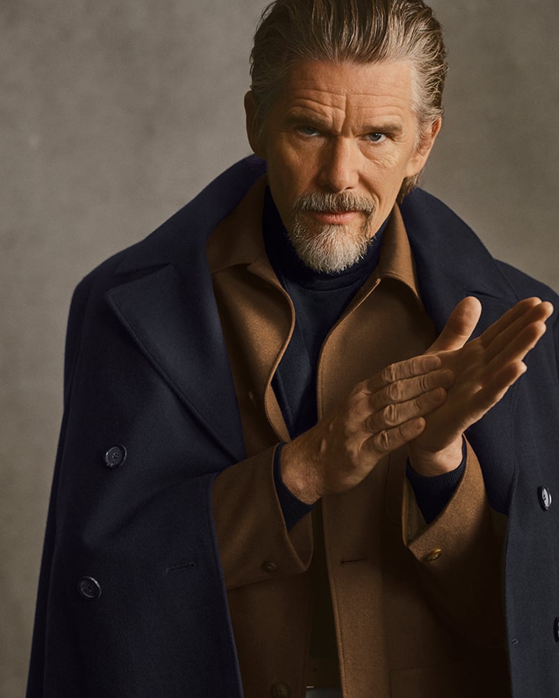 Ethan Hawke clasps his hands together in a navy overcoat draped over a camel jacket for Canali's fall-winter 2026 campaign.